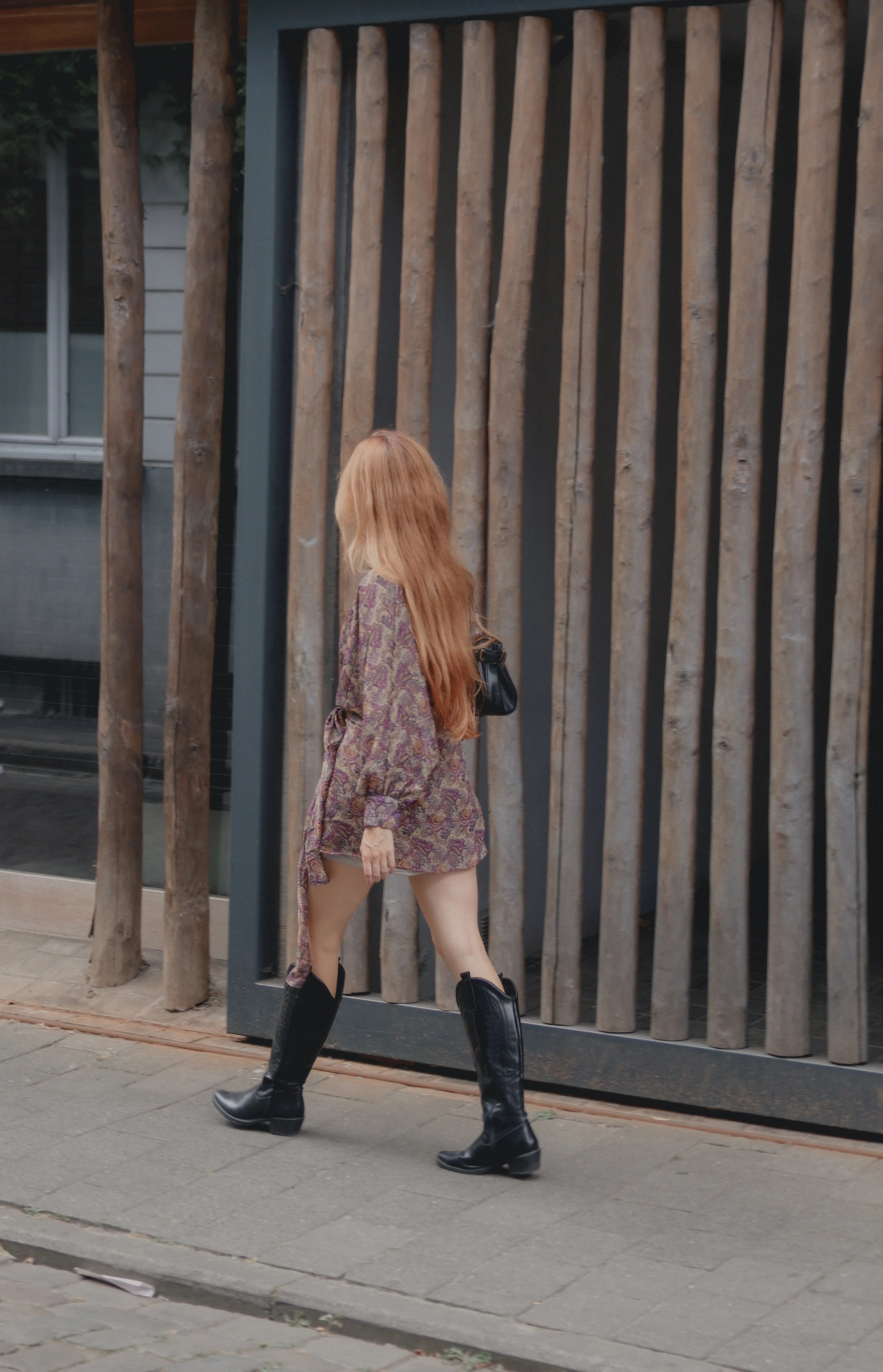 A woman with long red hair walks away from the camera on a sidewalk, wearing a short floral-patterned dress and tall black boots, in front of a modern wooden slat wall.