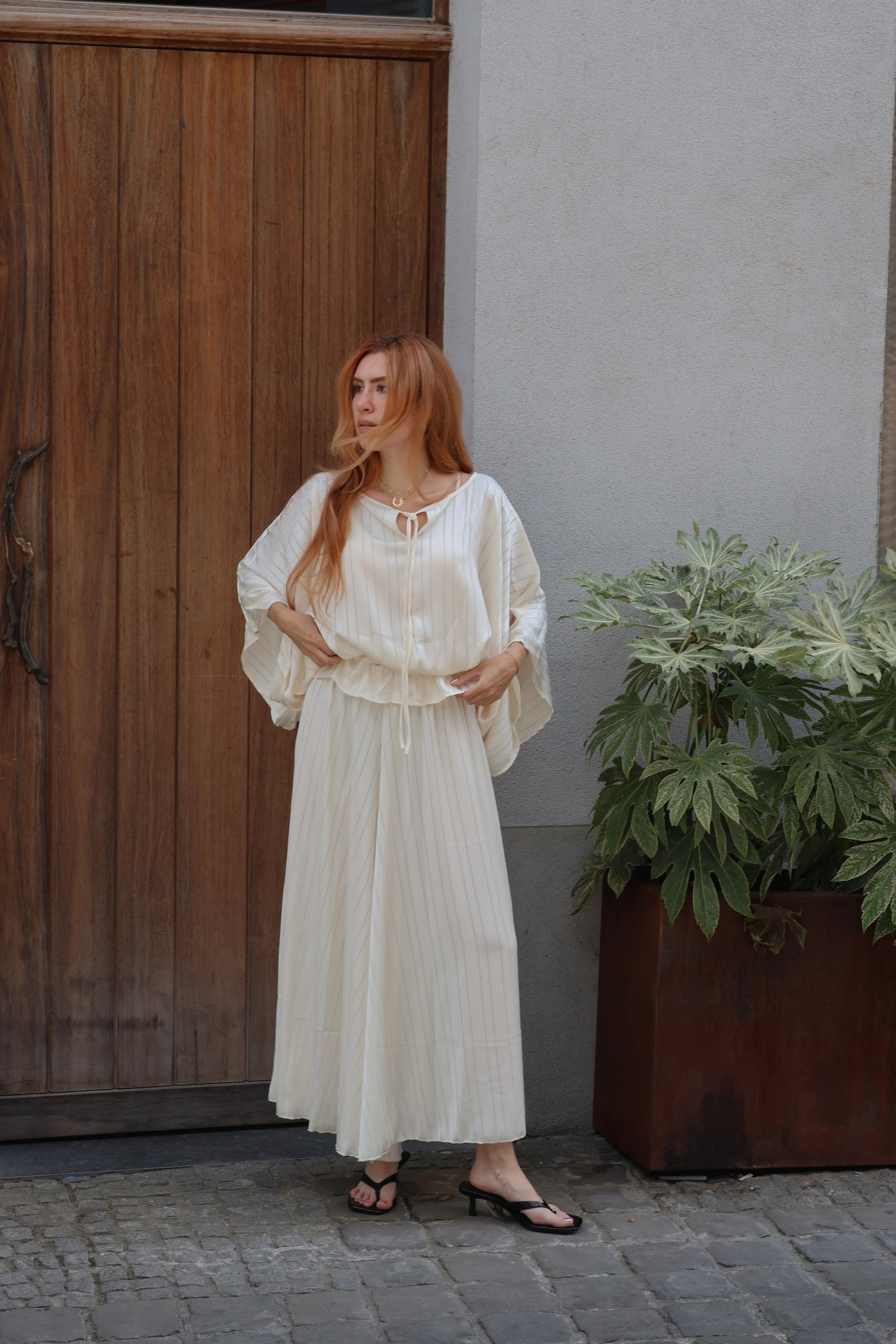 A woman with red hair wearing a cream-colored bohemian-style maxi dress with pleated details and wide sleeves, standing outdoors next to a wooden door and a potted plant.