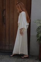 A woman with long red hair stands in profile wearing a loose, flowing white maxi dress with wide sleeves and black flip-flop sandals, standing outdoors next to a dark wooden door and a gray wall.