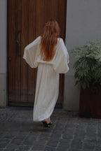 A woman with long red hair is seen from behind wearing a flowing cream-colored maxi dress with voluminous sleeves, standing on a cobblestone street in front of a rustic wooden door.