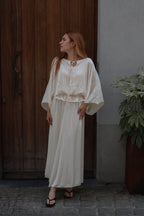 A woman with red hair wearing a cream-colored, long-sleeved pleated maxi dress and black sandals, standing in front of a dark wooden door and a gray wall with a potted plant.