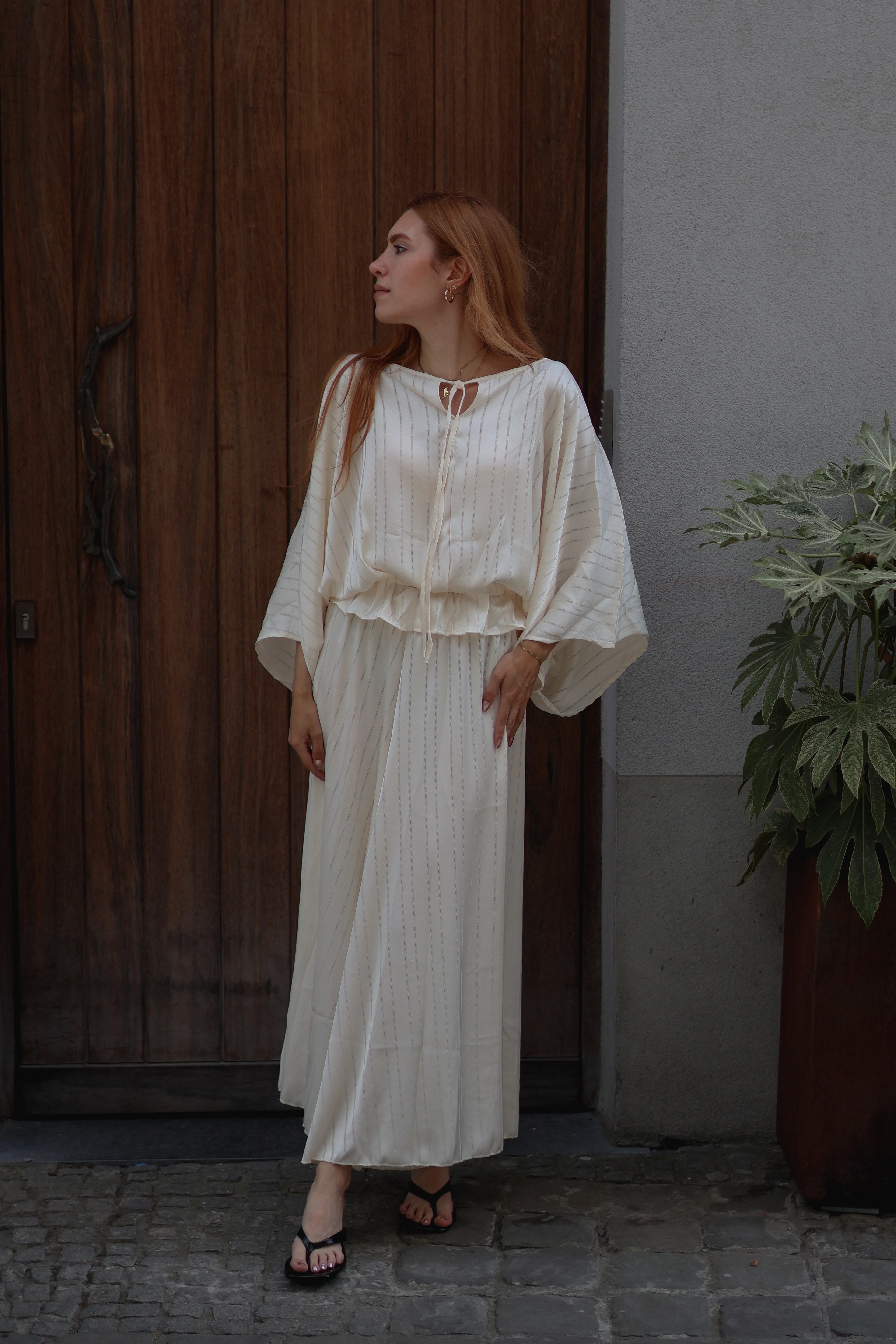 A woman with red hair wearing a cream-colored, long-sleeved pleated maxi dress and black sandals, standing in front of a dark wooden door and a gray wall with a potted plant.