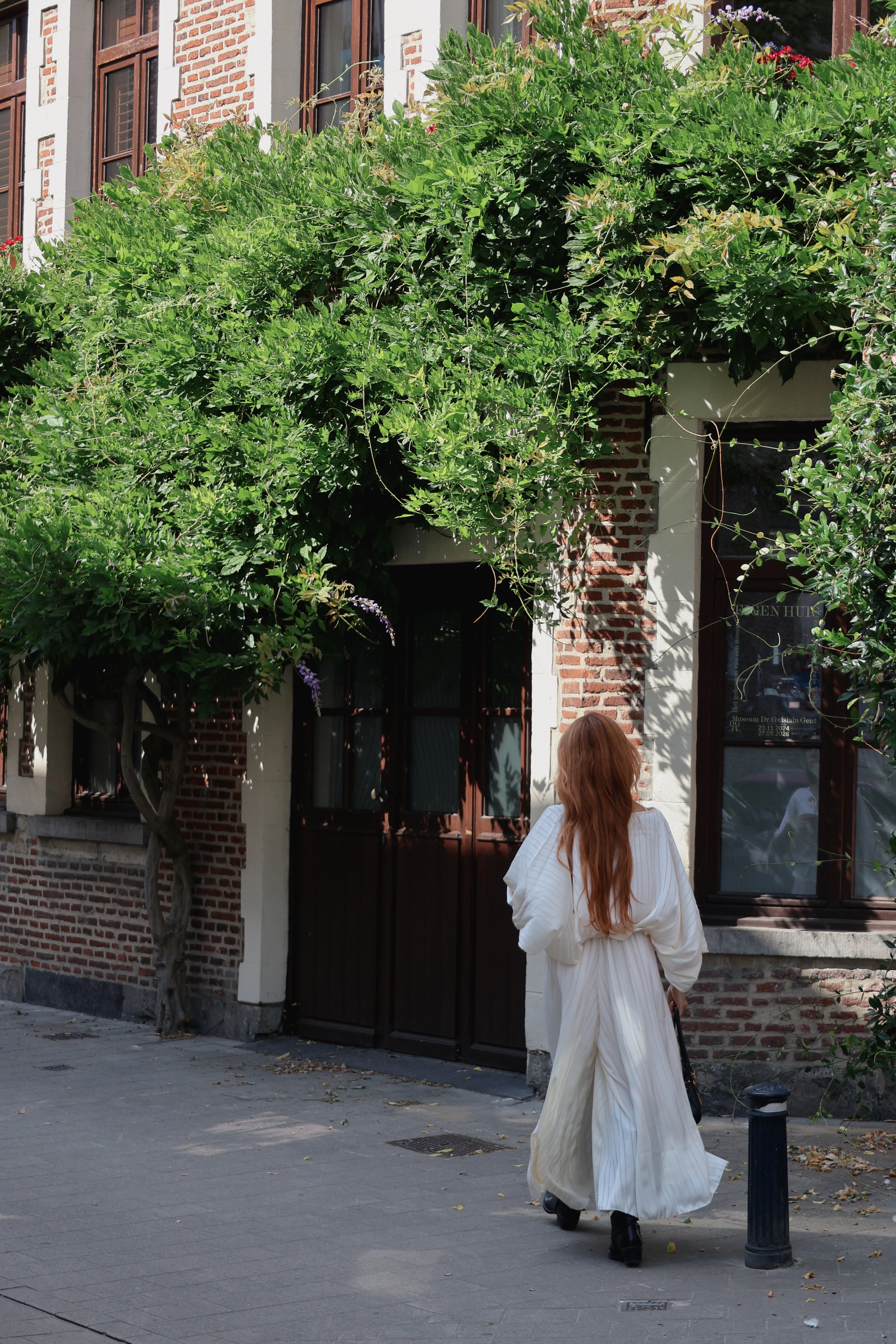 A woman with long red hair wearing a flowing white dress walks away from the camera on a sidewalk in front of a brick building with green vines covering the facade.