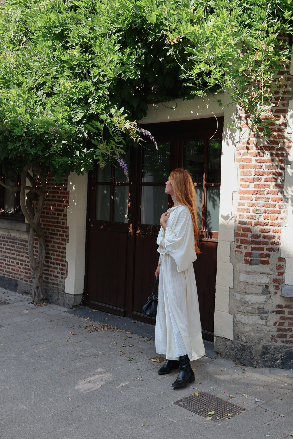 A woman with long red hair stands on a paved street in front of a brick building with a dark wooden door and lush green ivy. She is wearing a long-sleeved white maxi dress with a draped, flowing silhouette and black ankle boots.