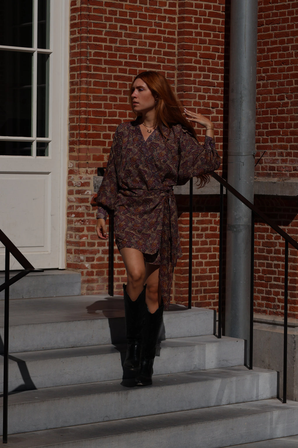 A woman with red hair walks down concrete steps wearing a dark patterned wrap dress with long sleeves and black knee-high boots, set against a brick building background.