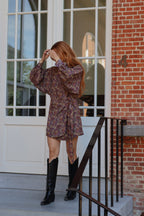 A woman with long red hair stands on a porch wearing a short, long-sleeved floral mini dress with a tie waist and black knee-high boots. The background features a white paneled door, brick wall, and a black metal railing.