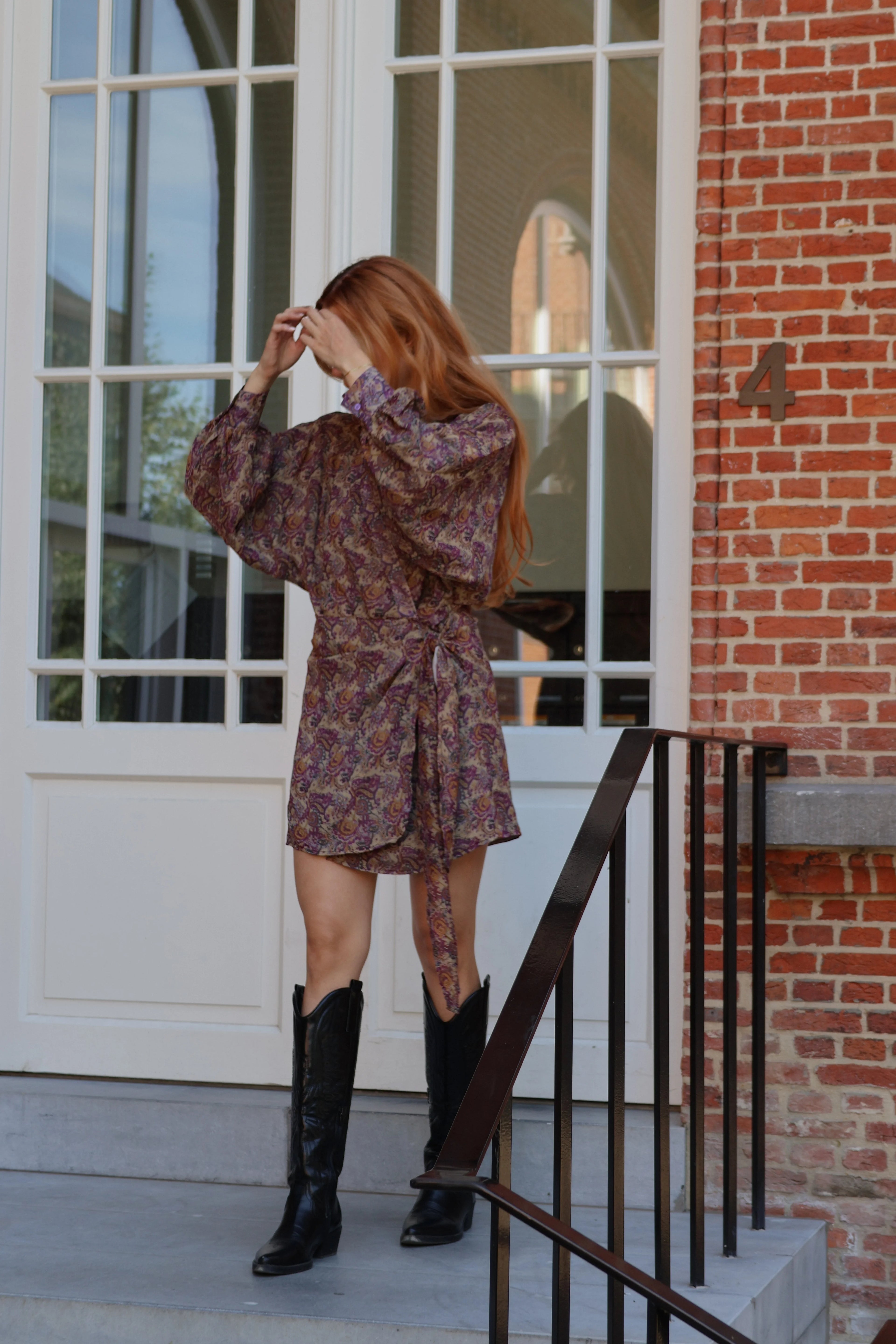A woman with long red hair stands on a porch wearing a short, long-sleeved floral mini dress with a tie waist and black knee-high boots. The background features a white paneled door, brick wall, and a black metal railing.
