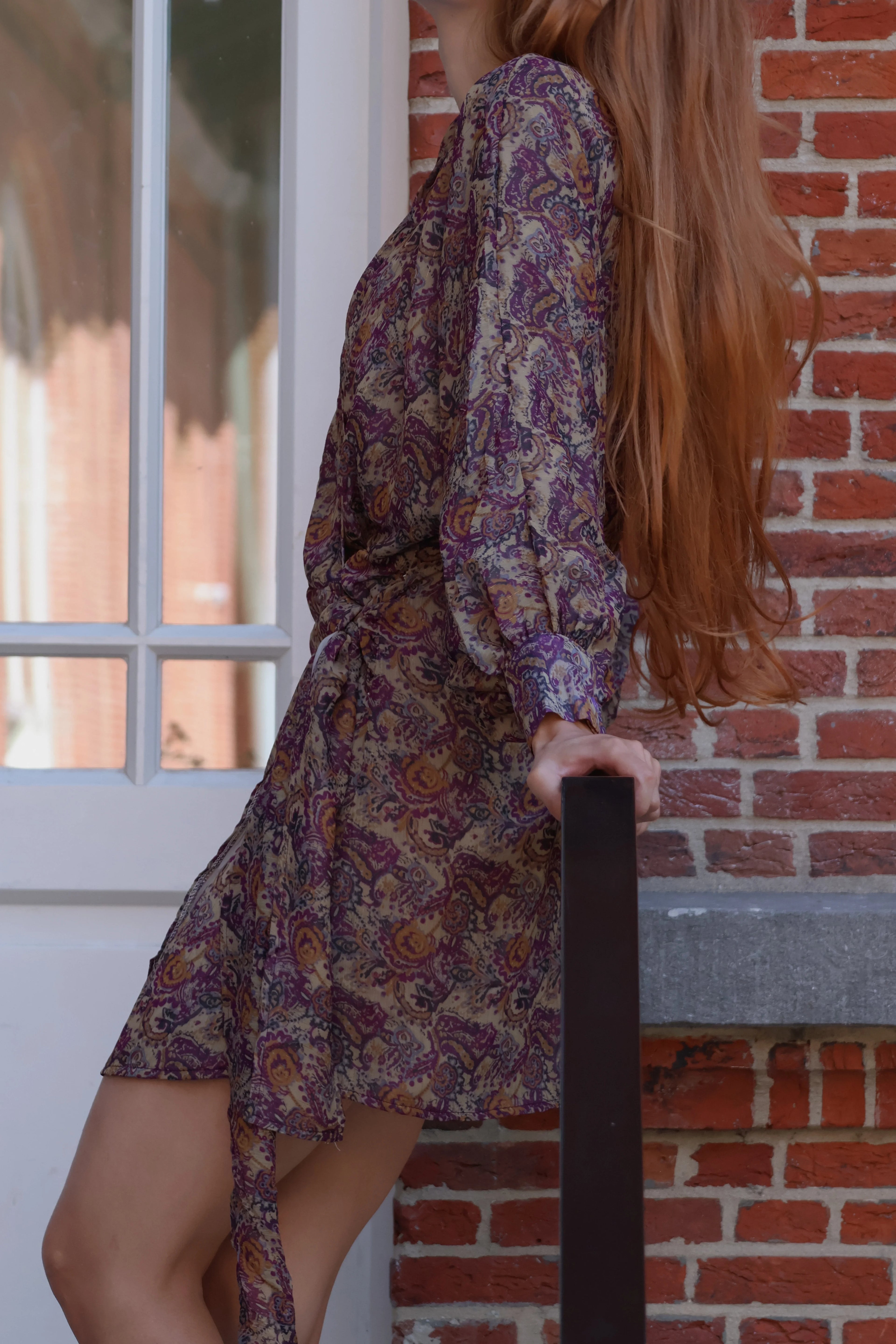 woman with red hair leaning against a stair railing and wearing a wrap mini dress with paisley print