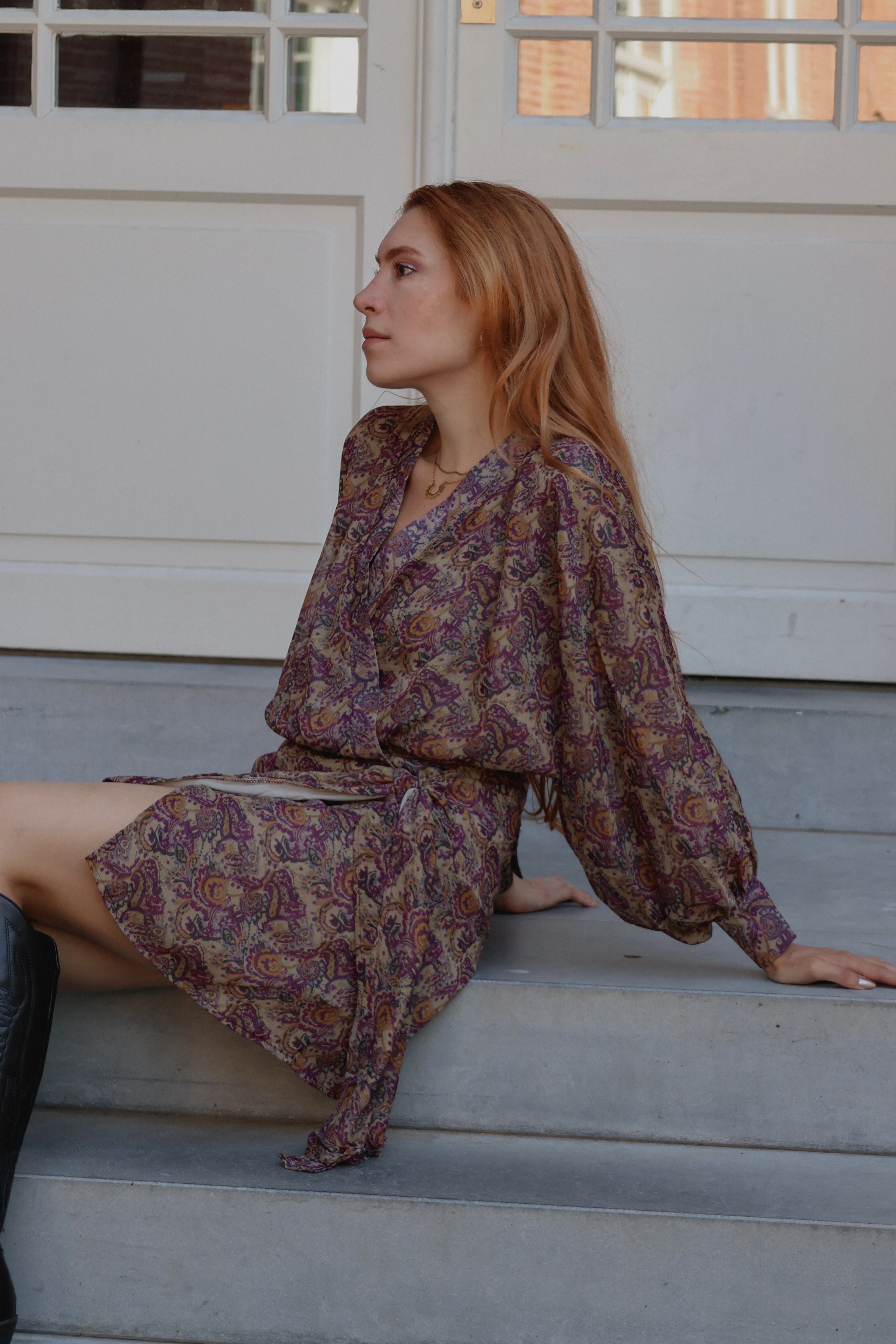 A woman with long light brown hair sits on concrete steps wearing a long-sleeved, knee-length bohemian paisley dress in purple, brown, and beige tones. She is posed in profile against a white paneled wall.