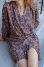 A woman wearing a long-sleeve wrap-style mini dress with a dense paisley pattern in purple, beige, and brown tones, sitting on outdoor concrete steps.