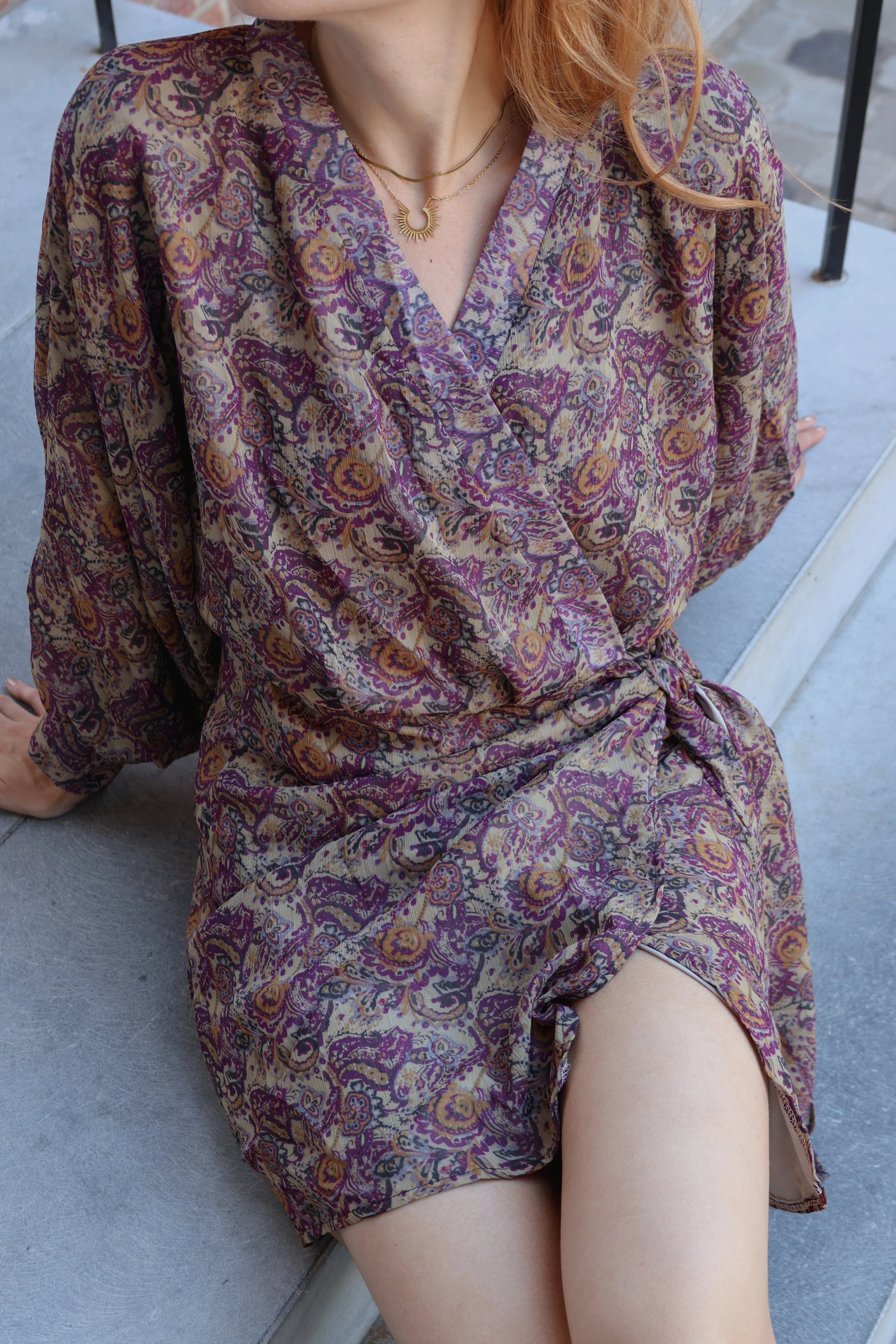 A woman wearing a long-sleeve wrap-style mini dress with a dense paisley pattern in purple, beige, and brown tones, sitting on outdoor concrete steps.