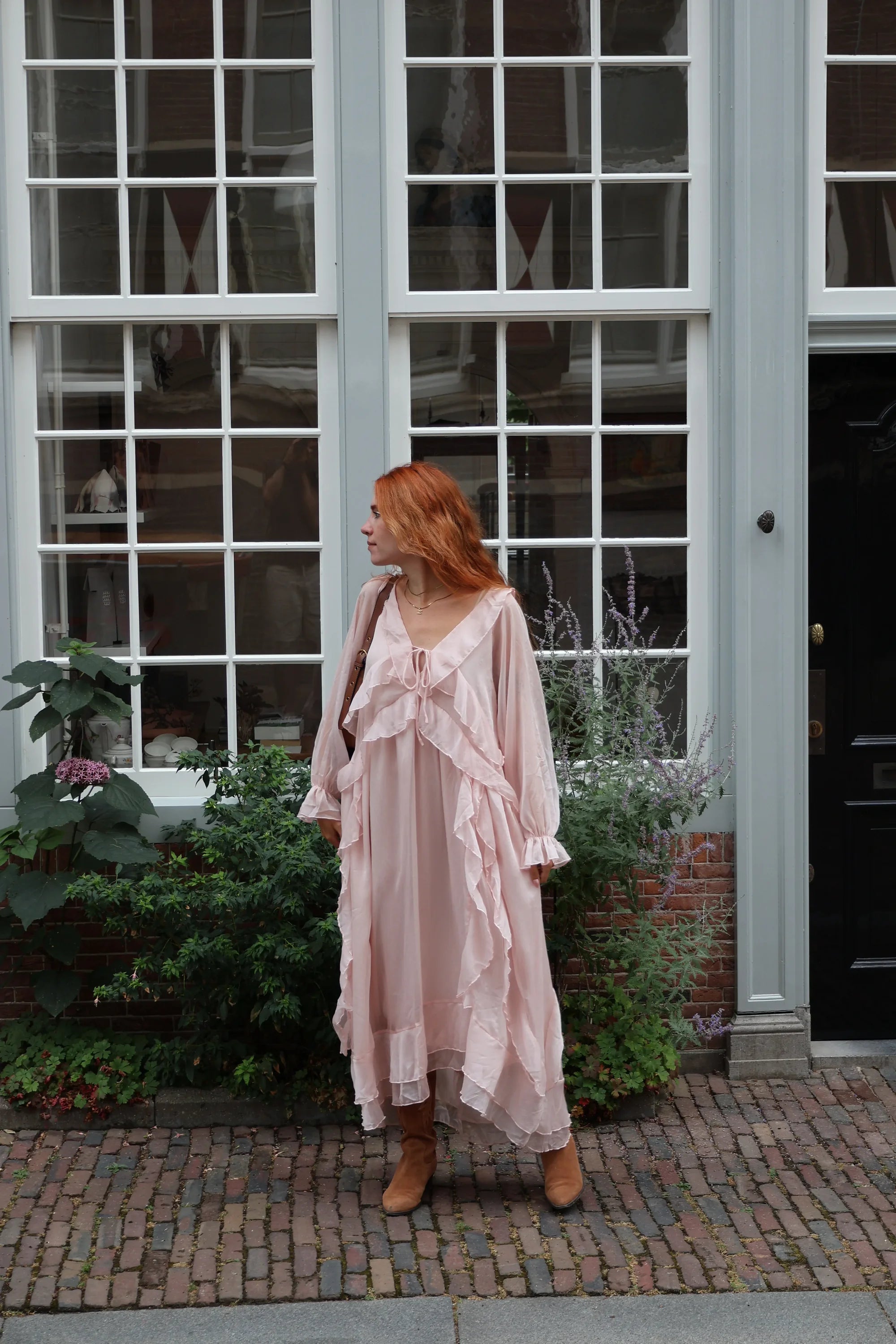 A woman with red hair stands on a brick sidewalk in front of a large white-framed window, wearing a light pink ruffled midi dress with a bow at the neckline and tan ankle boots.