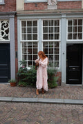 A woman with red hair stands on a cobblestone street in front of a classic brick building with grey-framed windows, wearing a long, light pink tiered maxi dress and holding a brown leather bag.