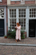 A woman with red hair stands on a cobblestone street in front of a classic brick building with grey-framed windows, wearing a long, light pink tiered maxi dress and holding a brown leather bag.