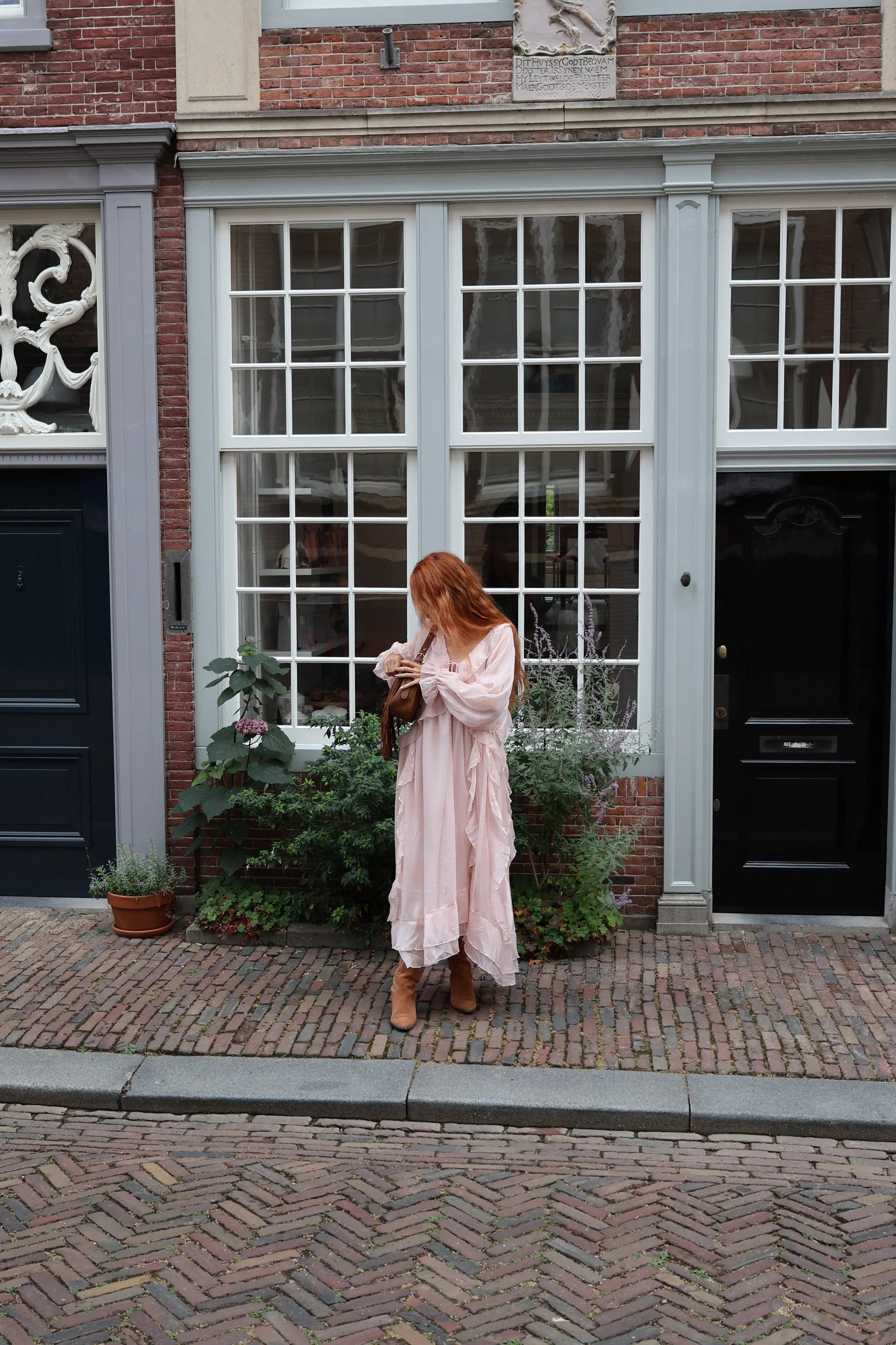 A woman with red hair stands on a cobblestone street in front of a classic brick building with grey-framed windows, wearing a long, light pink tiered maxi dress and holding a brown leather bag.