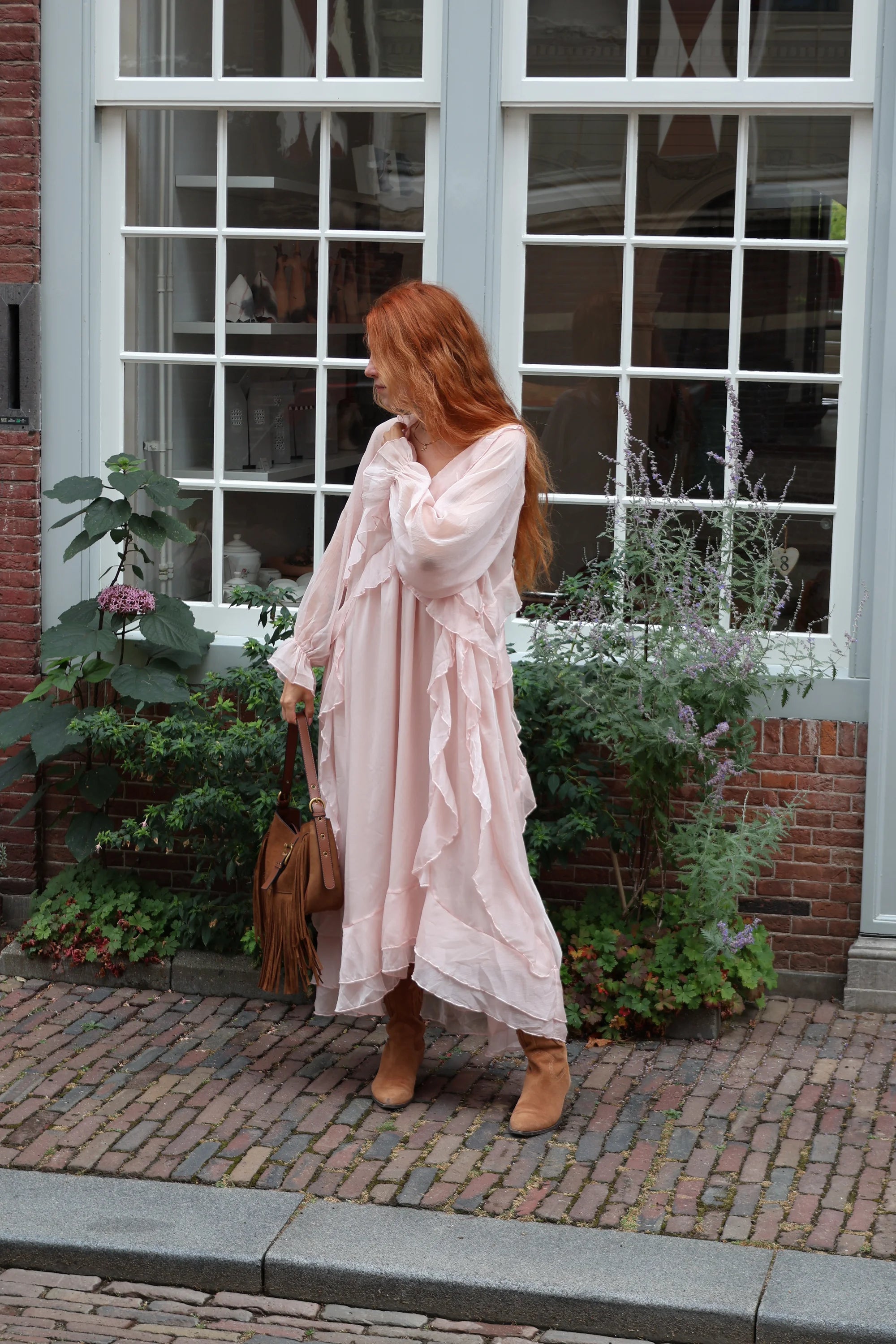 A woman with long red hair stands on a cobblestone street in front of a building with large white-framed windows and plants. She is wearing a flowing light pink long-sleeve maxi dress with ruffled layers and tan ankle boots, and holding a brown fringe handbag.