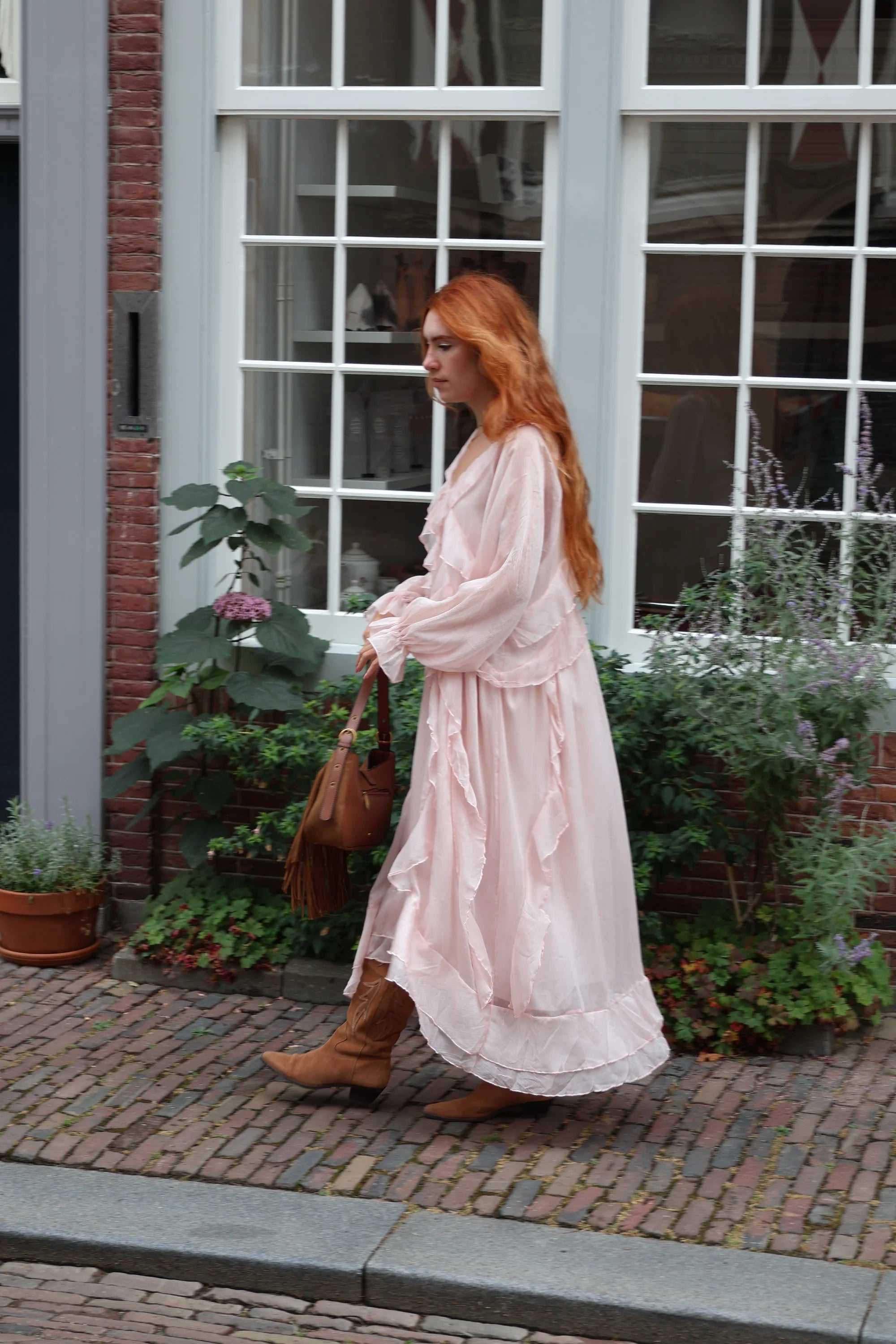 A woman with long red hair walks along a cobblestone street wearing a light pink long-sleeve maxi dress, brown boots, and carrying a brown leather handbag.