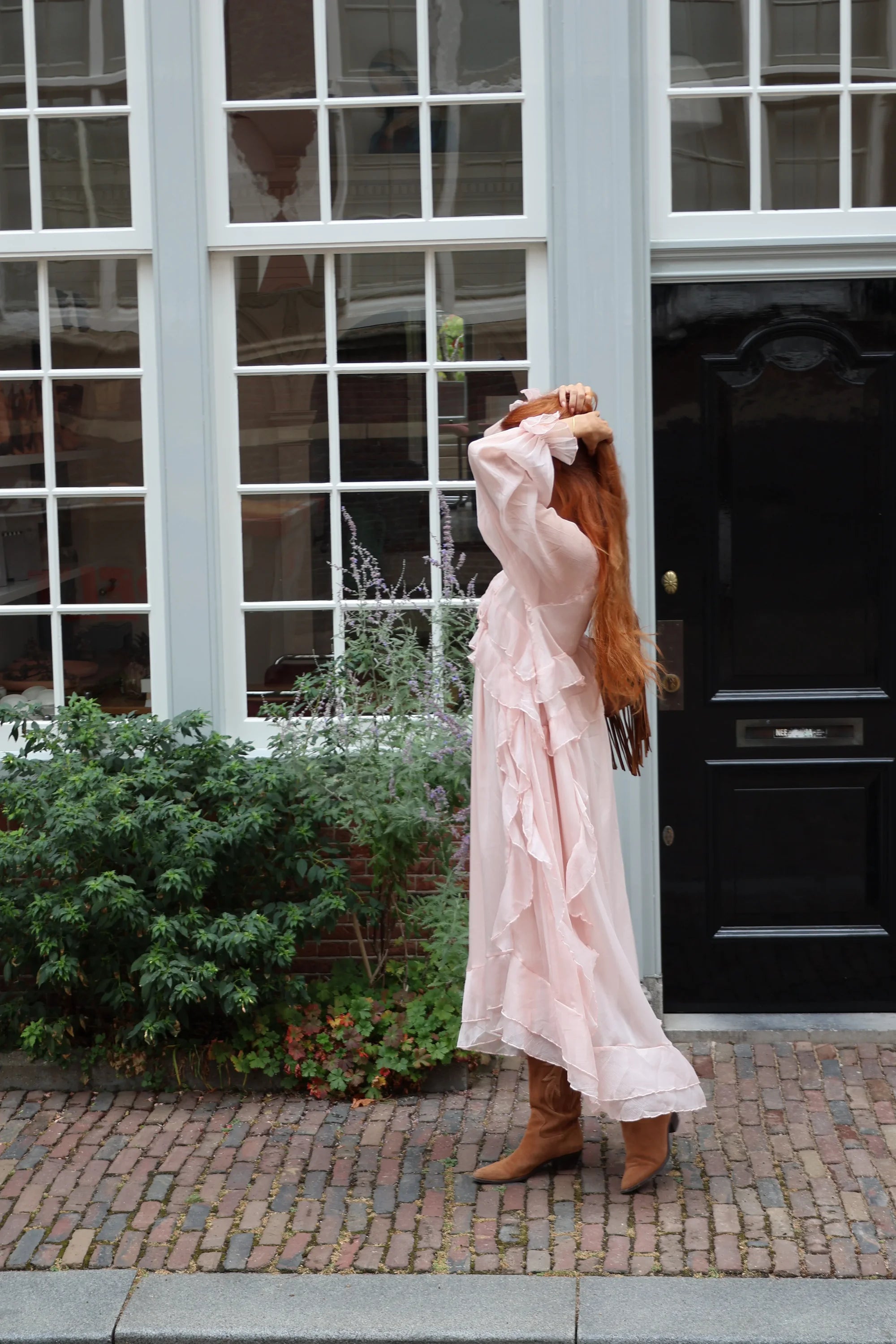 A woman with long light brown hair stands on a cobblestone street in front of a white building with large windows and a black door, wearing a long, light pink ruffled maxi dress and brown ankle boots.