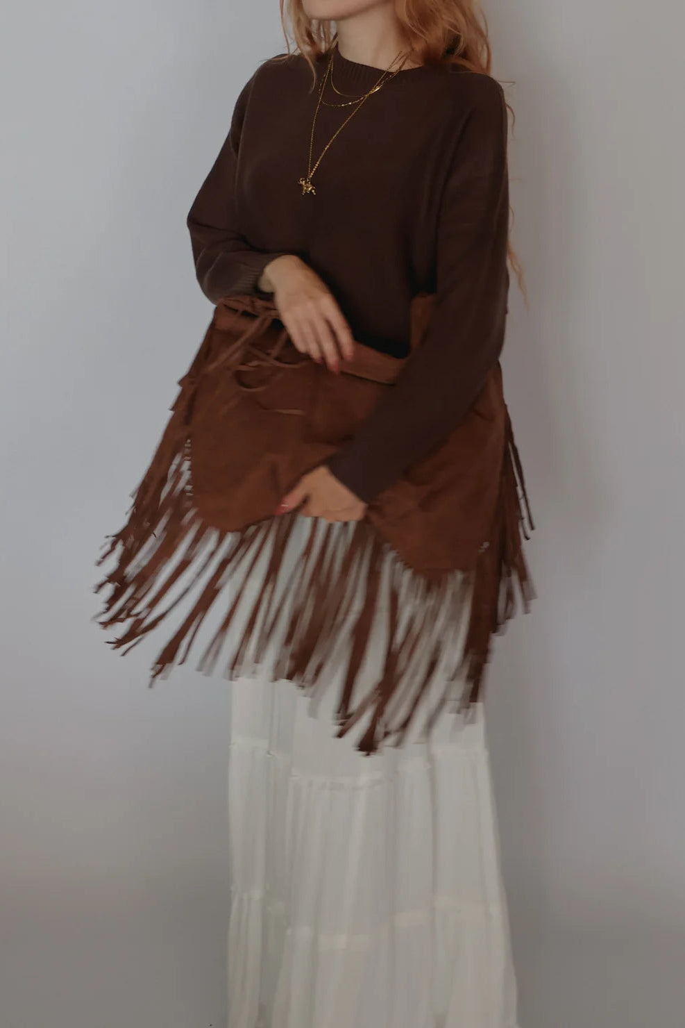 A person wearing a dark brown long-sleeve top and a matching brown fringe bag, paired with a white tiered skirt, against a plain light gray background.