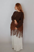 A woman with red hair stands in profile wearing a dark brown long-sleeve top, a matching brown fringed wrap or skirt overlay, and a long white tiered skirt, against a plain light gray studio background.