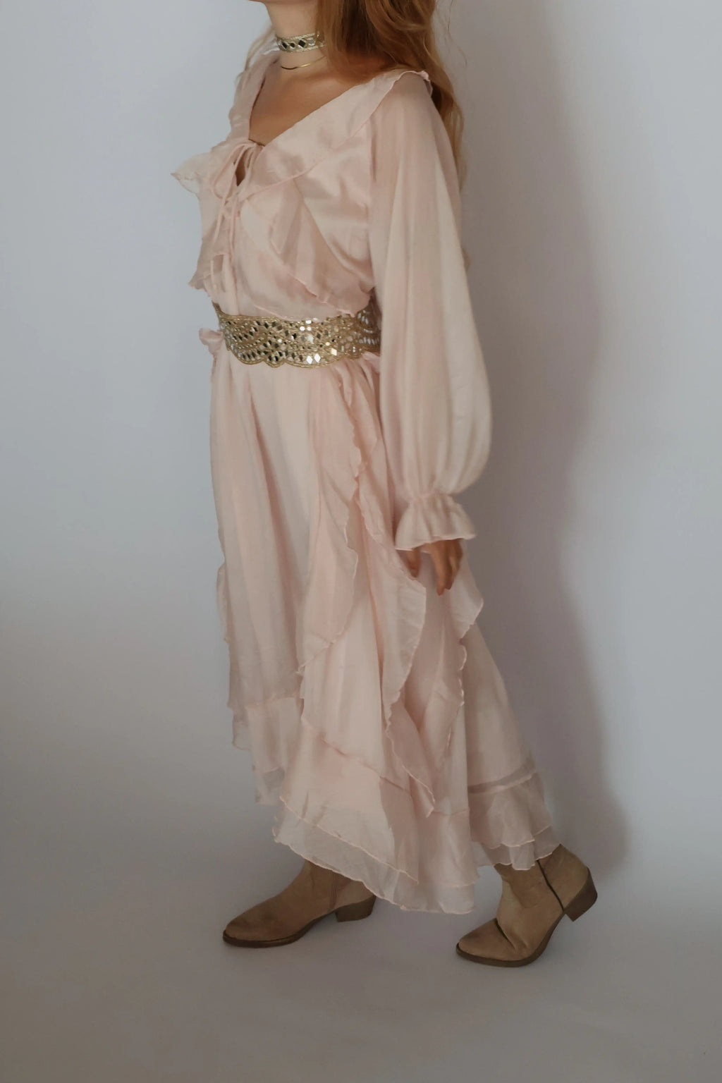 A woman wearing a light blush pink bohemian-style maxi dress with a wide, embellished gold sequin waist belt, paired with beige flat shoes against a plain light gray background.