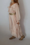 A woman wearing a light blush pink bohemian-style maxi dress with a wide, embellished gold sequin waist belt, paired with beige flat shoes against a plain light gray background.