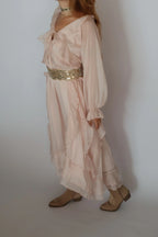 A woman wearing a light blush pink bohemian-style maxi dress with a wide, embellished gold sequin waist belt, paired with beige flat shoes against a plain light gray background.