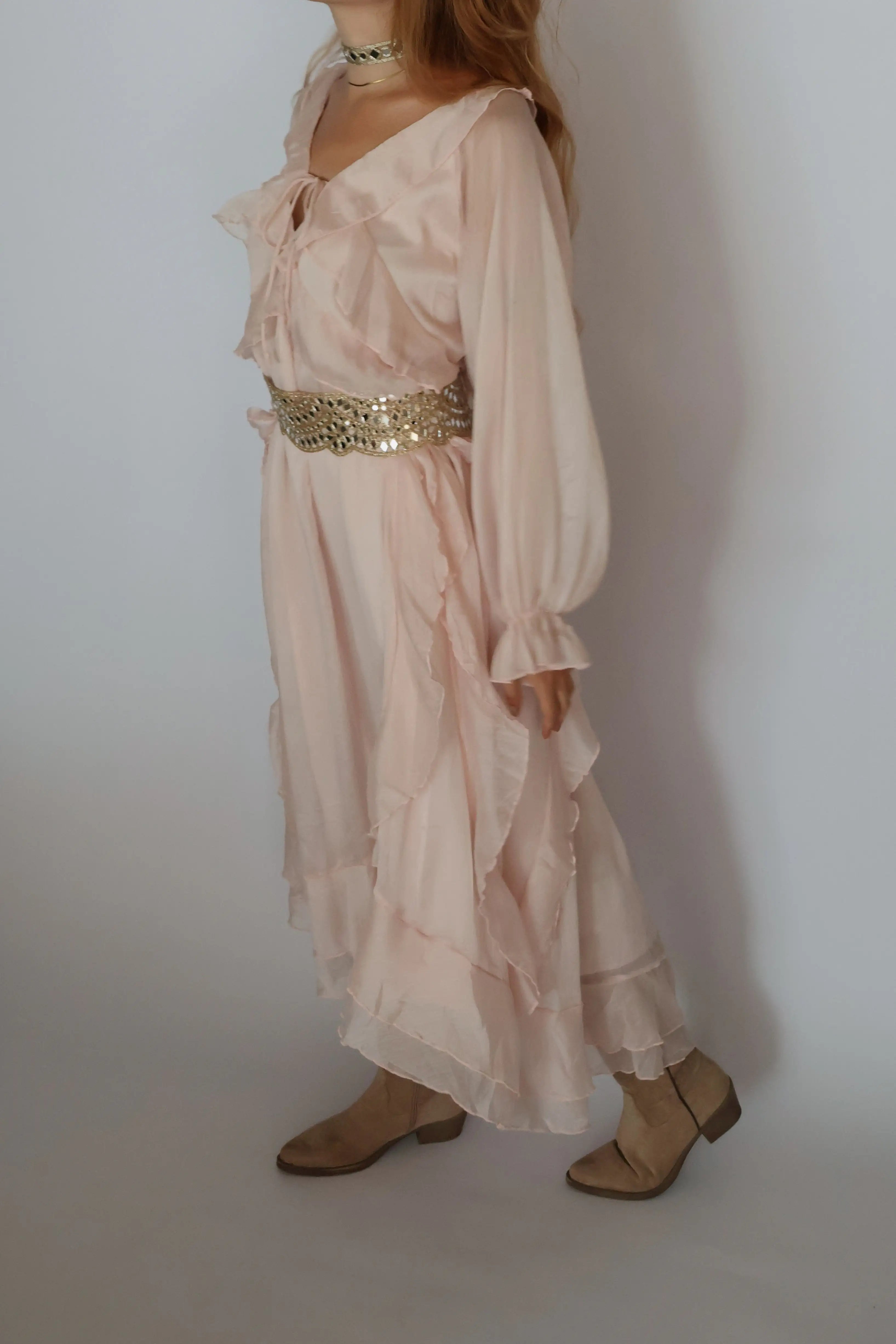 A woman wearing a light blush pink bohemian-style maxi dress with a wide, embellished gold sequin waist belt, paired with beige flat shoes against a plain light gray background.