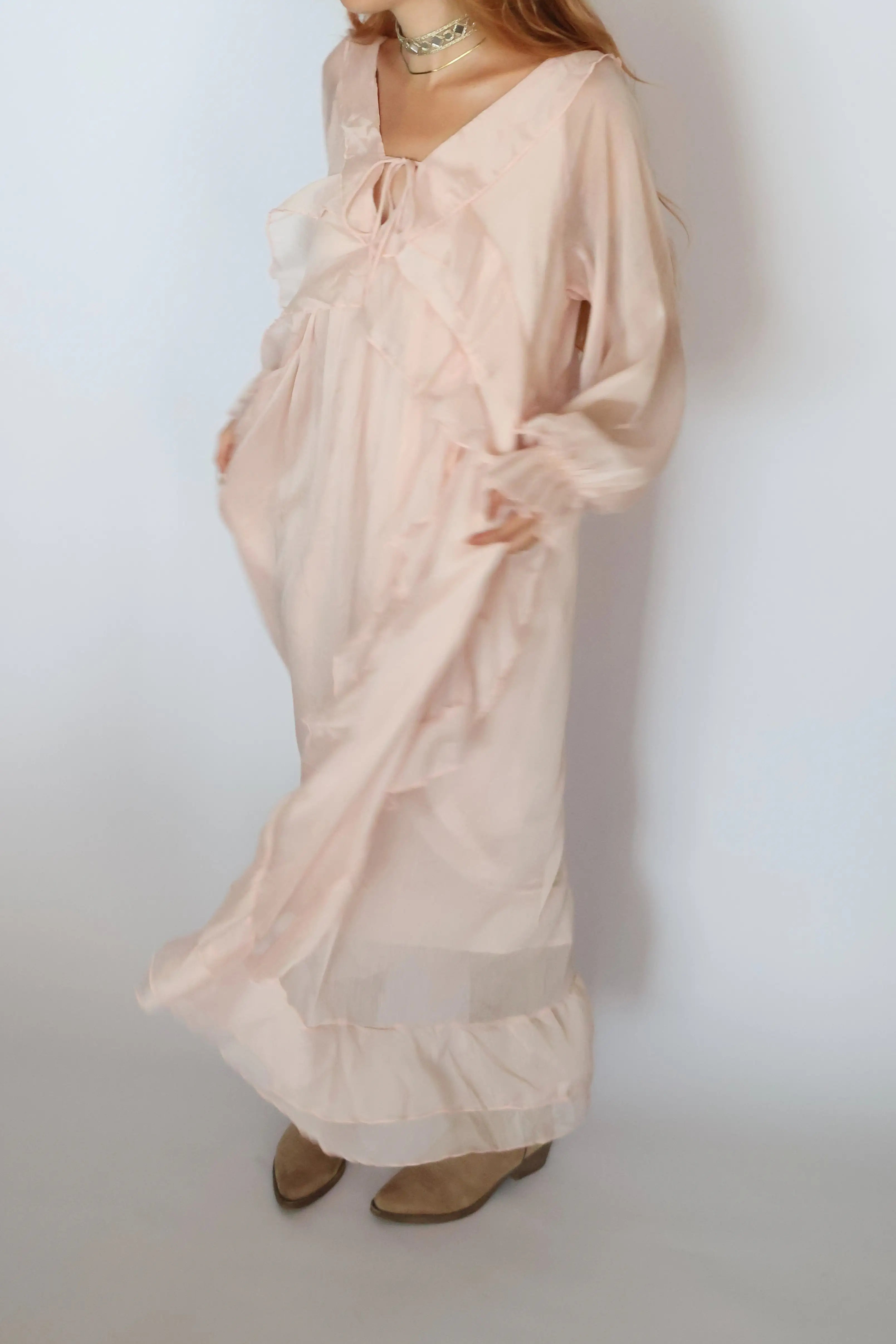A woman wearing a long, flowing light pink maxi dress with ruffles and sheer long sleeves, paired with tan shoes, standing against a plain white background.