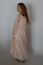 A woman with long wavy light brown hair is shown from the back wearing a pale blush pink, tiered maxi dress with sheer long sleeves and ruffled details, standing against a plain light gray studio background.