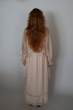 A woman with long, wavy red hair is shown from behind wearing a pale blush pink long-sleeved maxi dress with ruffled details, standing against a plain light gray studio background.