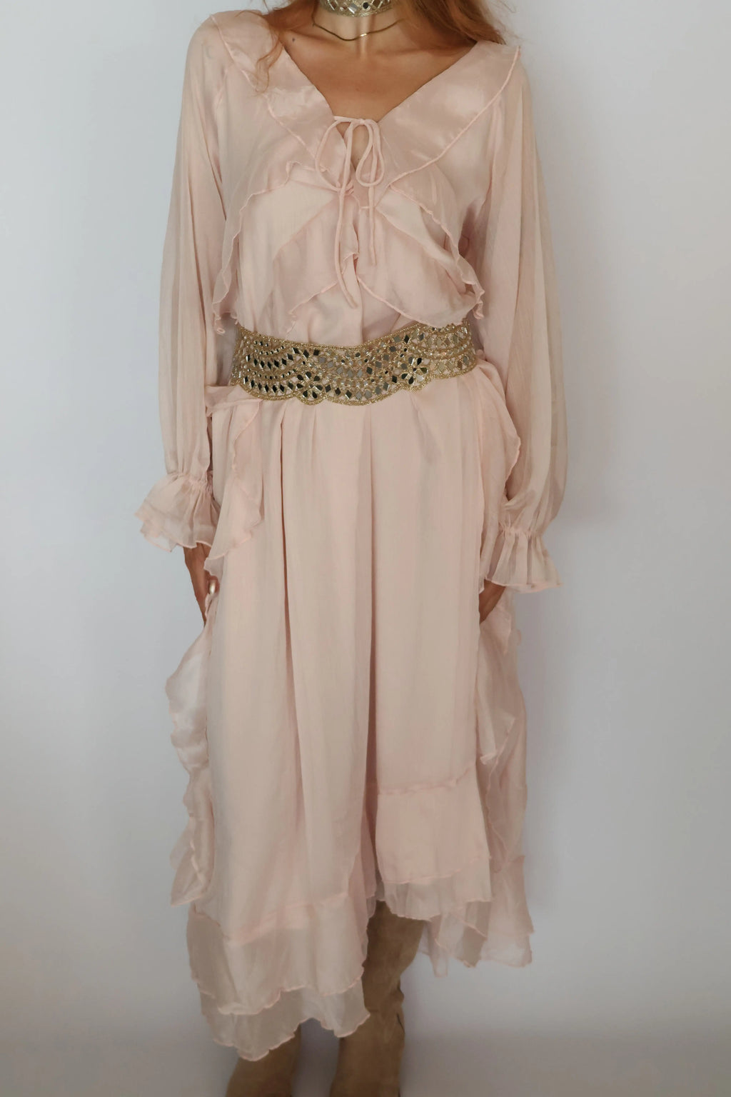 A woman wearing a light blush pink long-sleeve midi dress with a ruffled tie neckline, a wide embellished gold belt at the waist, and a layered ruffled hem, paired with tan boots against a plain white wall.
