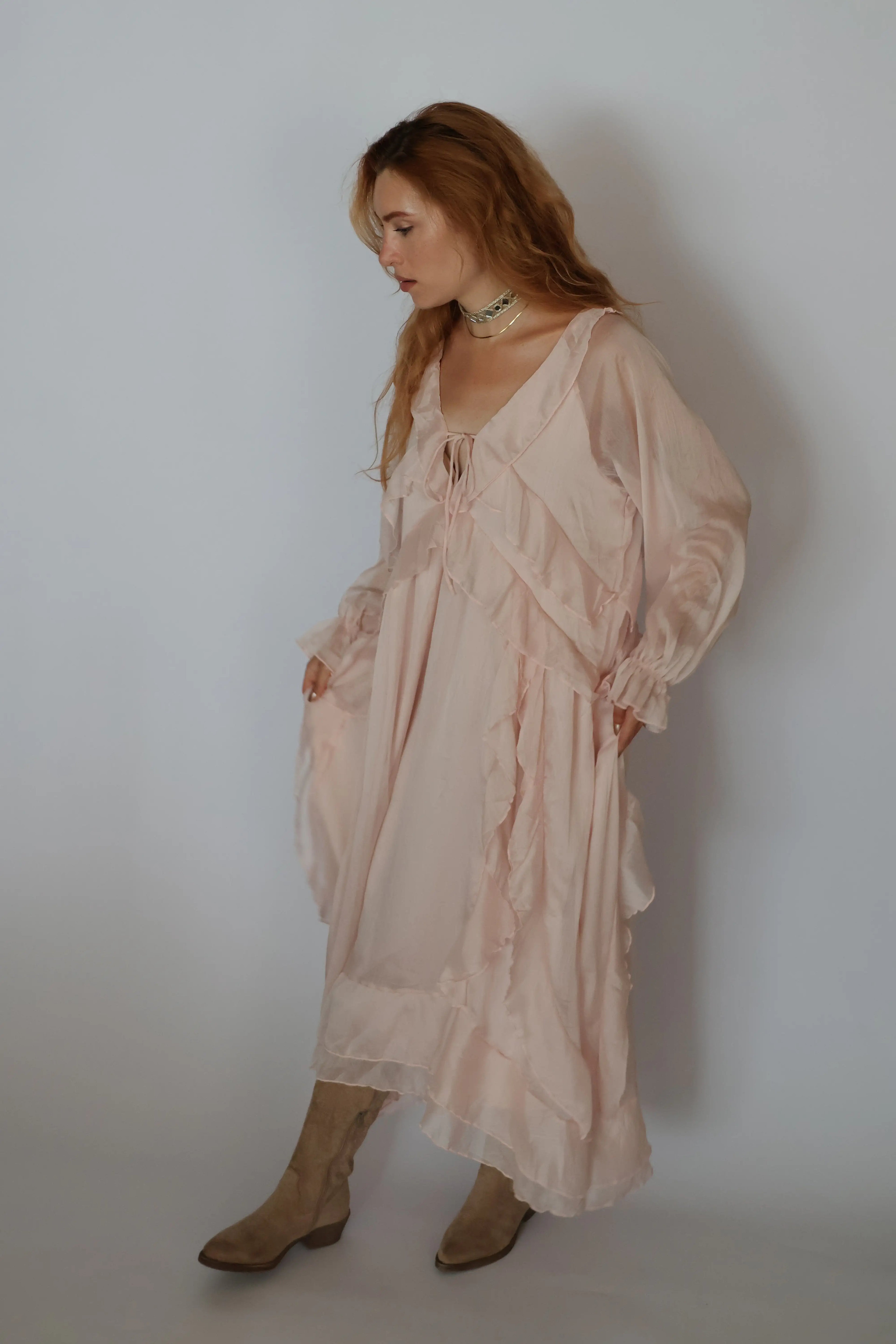 A woman wearing a long, flowing light pink sheer maxi dress with ruffles and long sleeves, paired with beige ankle boots, standing against a plain light gray wall.