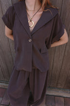 A woman wearing a dark brown short-sleeve button-front top with a wide collar and matching loose pants, standing against a wooden fence.