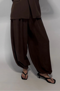 A woman takes a mirror selfie wearing a dark brown two-piece outfit consisting of a short-sleeve button-front top and matching loose harem-style pants, paired with black sandals against a plain light gray studio background.