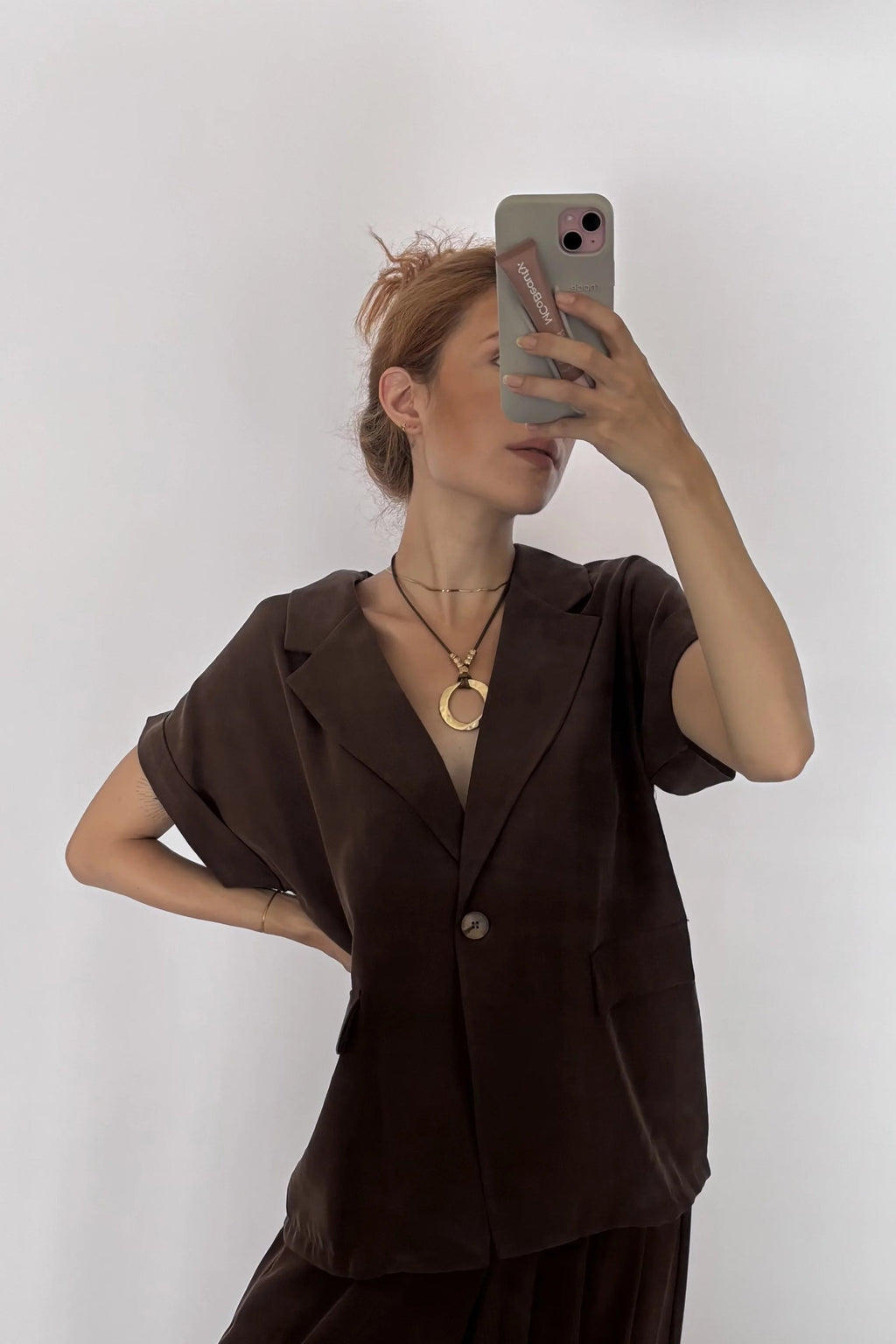 A woman takes a mirror selfie wearing a dark brown short-sleeve blazer and matching wide-leg trousers, paired with black sandals and gold necklaces, against a plain light gray background.