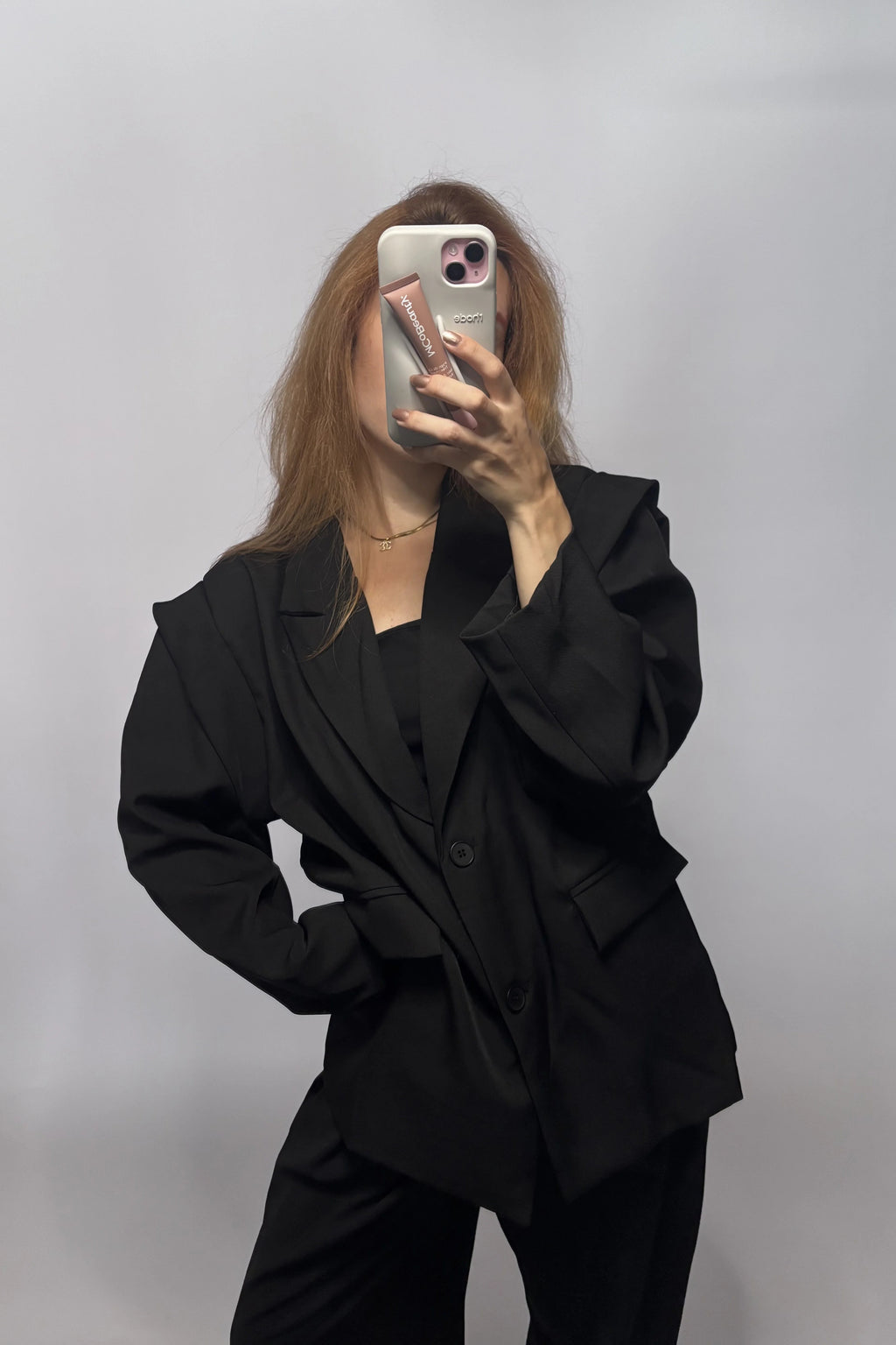 A woman takes a mirror selfie wearing an oversized black blazer and matching black pants against a plain light gray background.