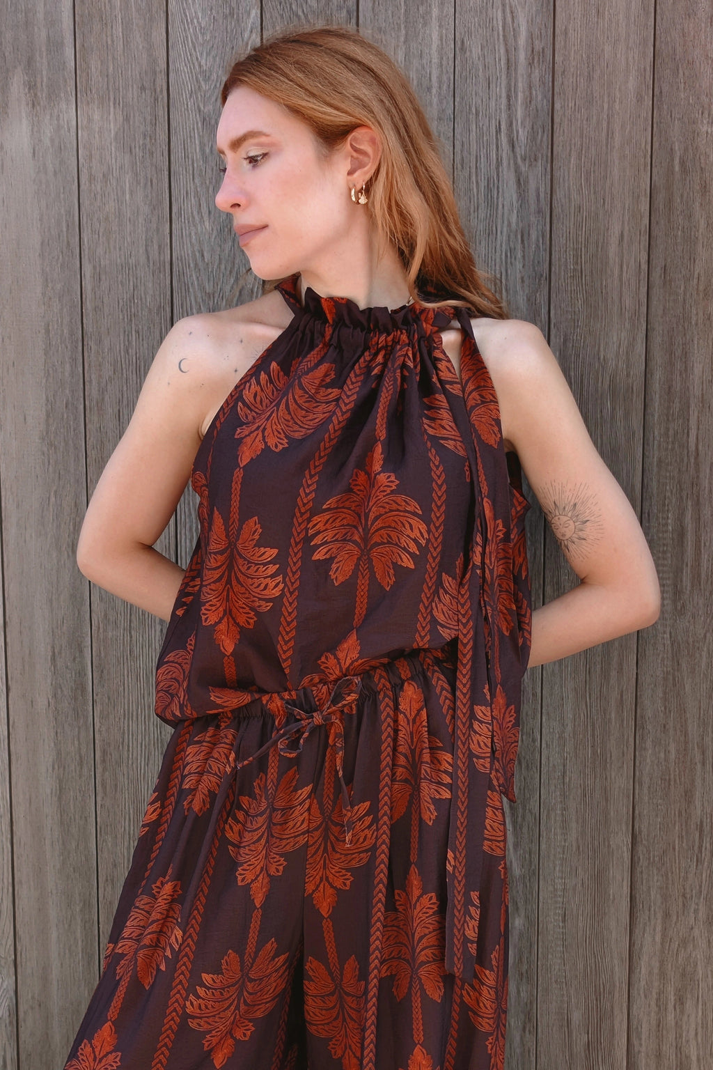 A woman wearing a dark brown halter jumpsuit with a rust-orange palm leaf pattern, standing against a weathered gray wooden plank wall.