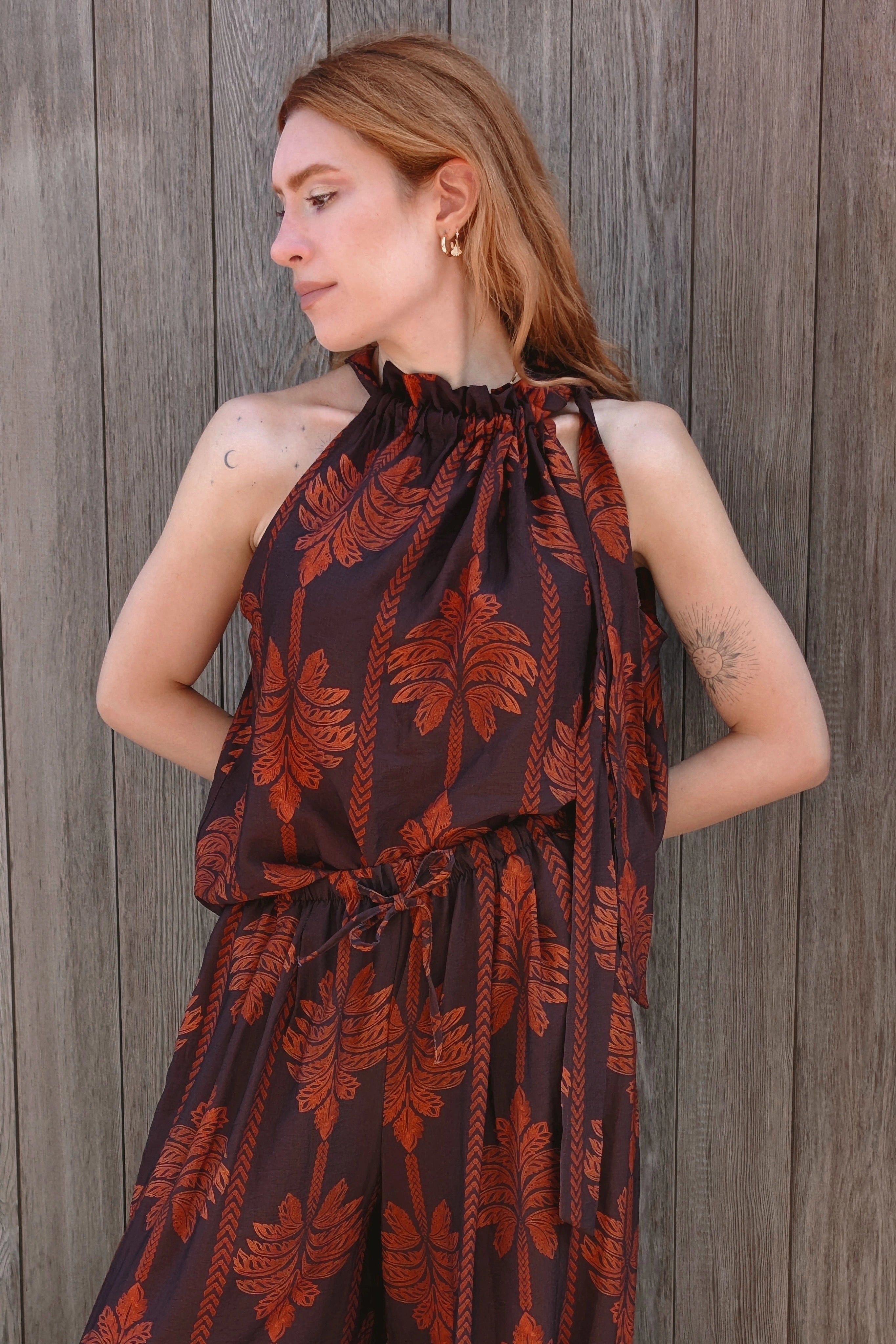A woman wearing a dark brown halter jumpsuit with a rust-orange palm leaf pattern, standing against a weathered gray wooden plank wall.