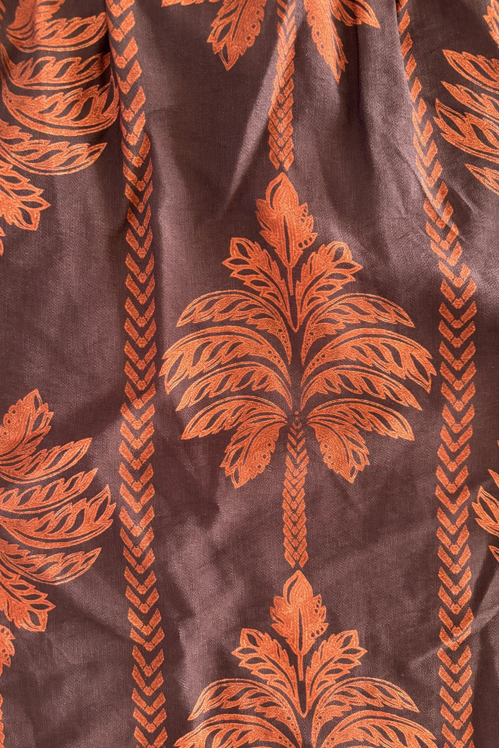 A close-up of a brown fabric with a repeating orange palm tree and leaf pattern, bordered by vertical orange chevron stripes.
