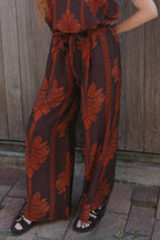 A woman wearing a brown and rust-orange patterned sleeveless jumpsuit with wide legs, standing outdoors against a weathered wooden fence.