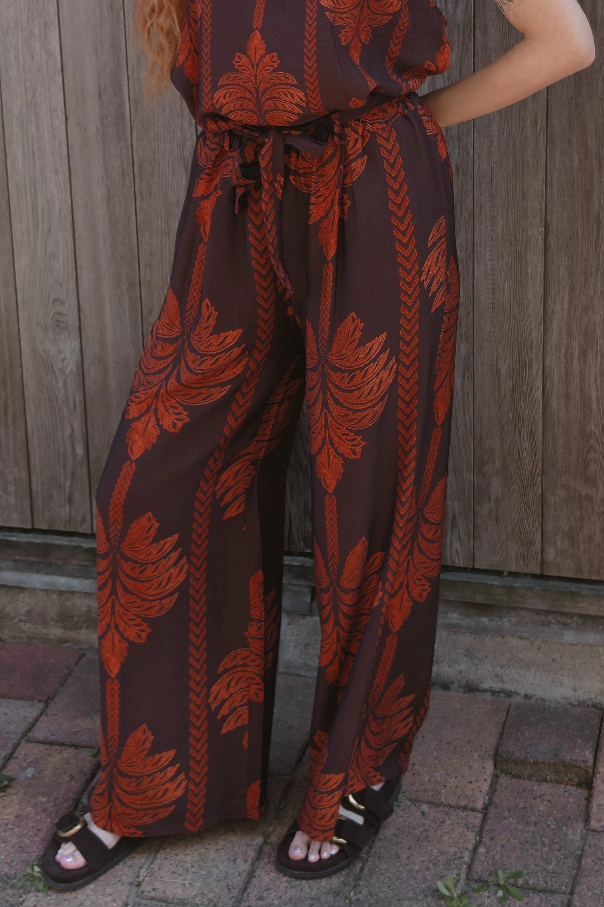 A woman wearing a brown and rust-orange patterned sleeveless jumpsuit with wide legs, standing outdoors against a weathered wooden fence.