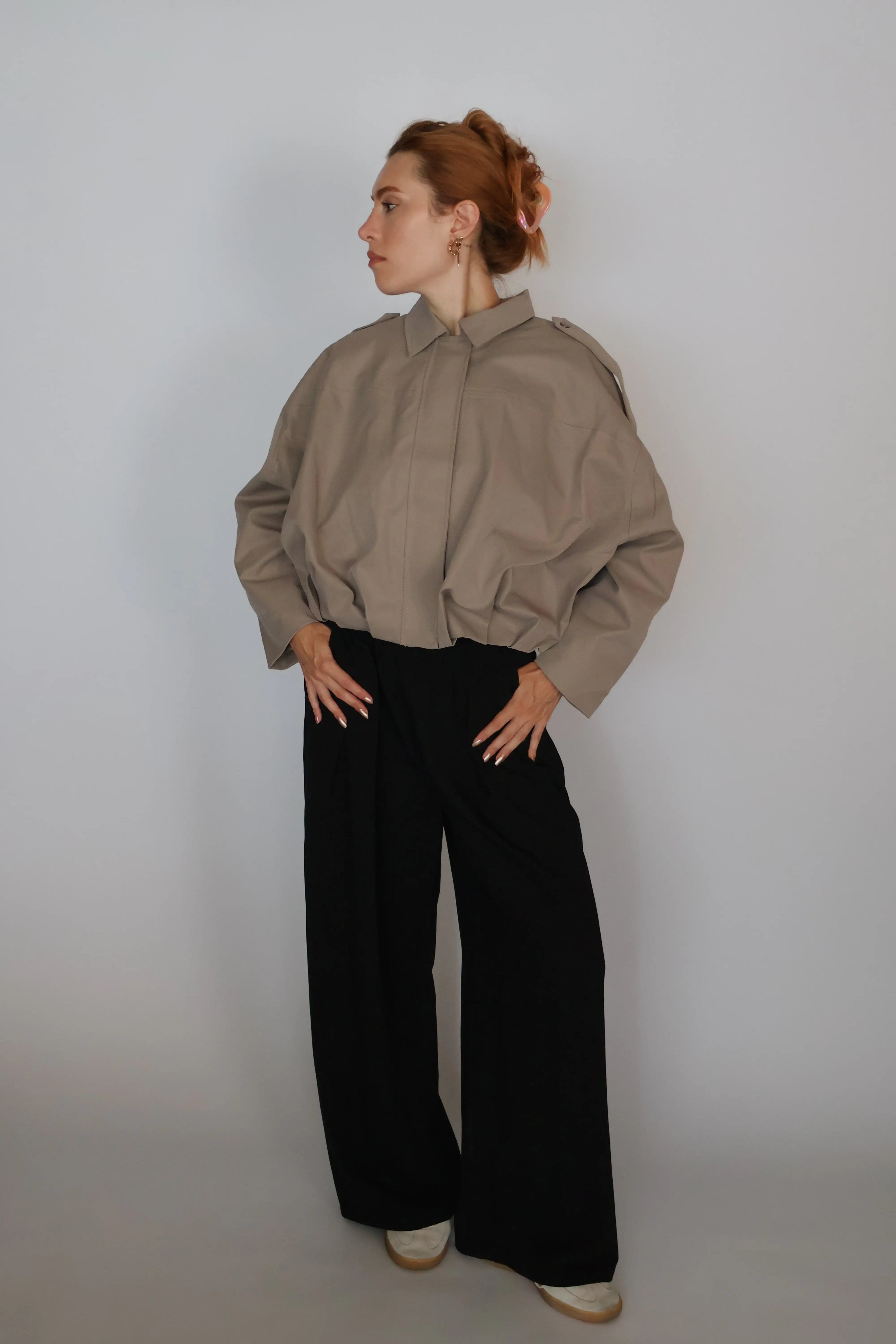 A woman with red hair tied up is wearing a loose taupe-brown long-sleeve button-up shirt and black wide-leg trousers, standing against a plain light gray studio background.
