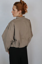 A woman with red hair in an updo is shown from the back wearing a loose taupe-beige cropped jacket with dramatic oversized sleeves and black pants, against a plain light gray background.