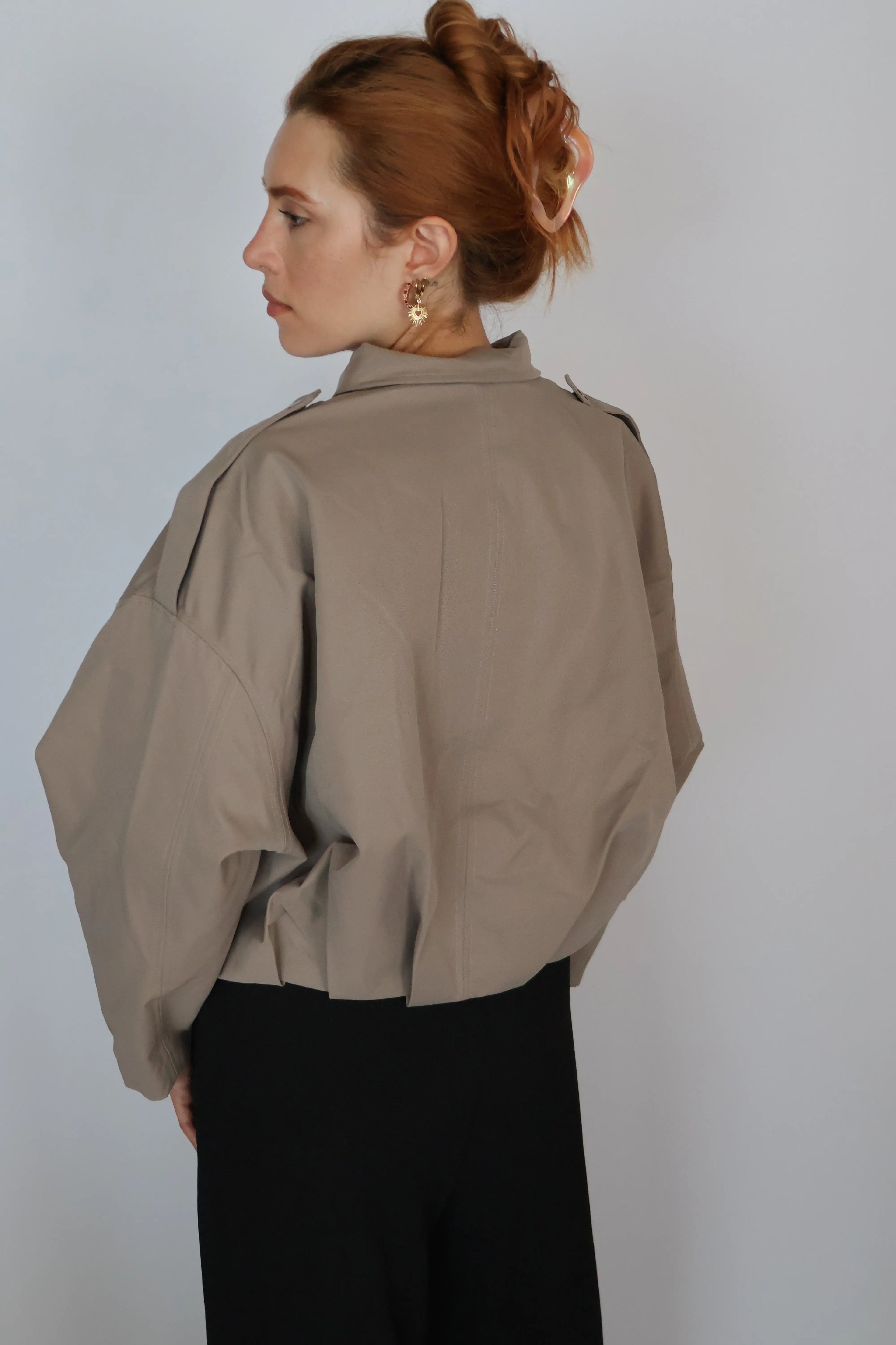 A woman with red hair in an updo is shown from the back wearing a loose taupe-beige cropped jacket with dramatic oversized sleeves and black pants, against a plain light gray background.