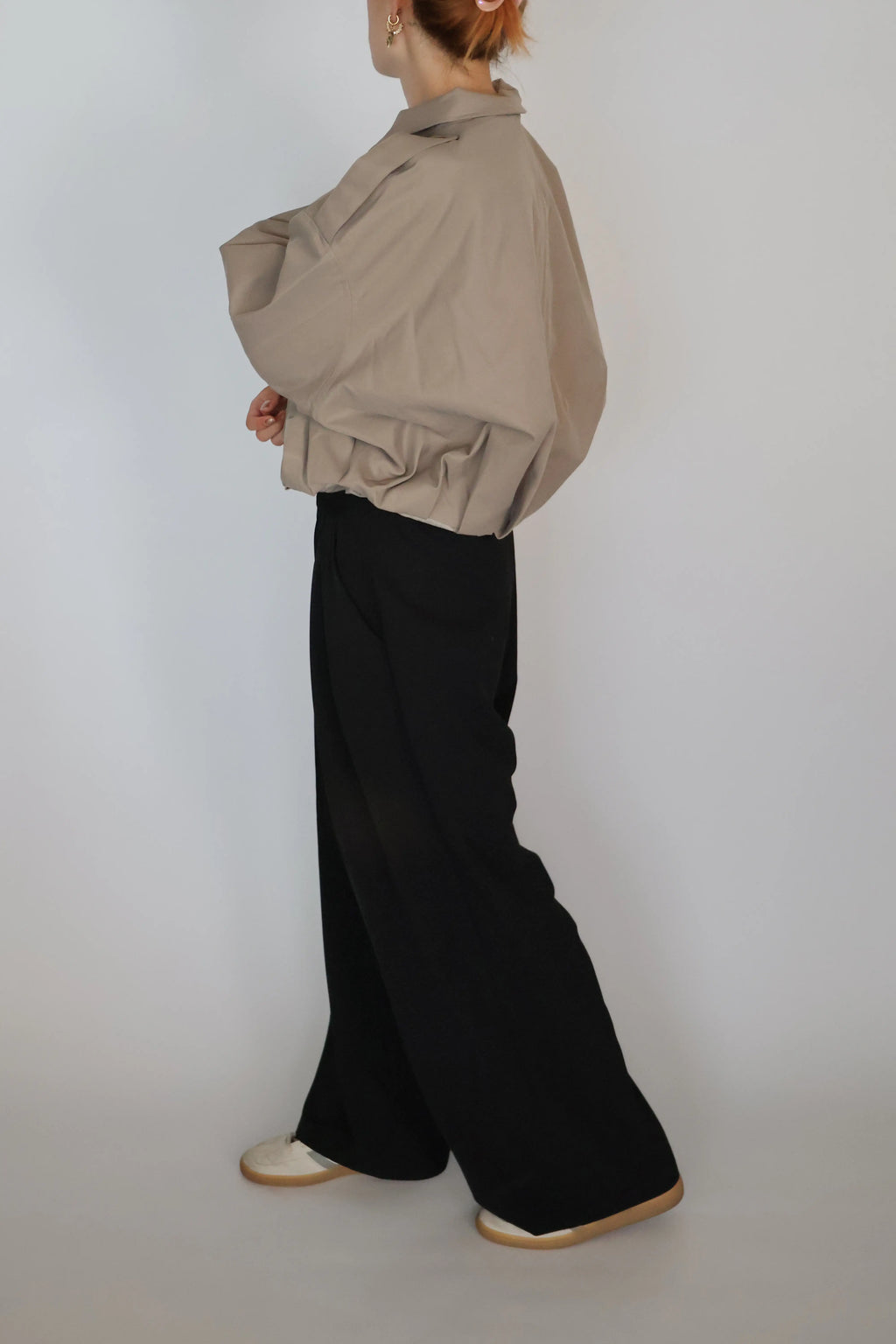 A model shown from the back wearing a beige oversized long-sleeve shirt and black wide-leg trousers, paired with white shoes, against a plain light gray studio background.