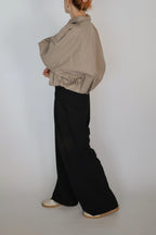 A model shown from the back wearing a beige oversized long-sleeve shirt and black wide-leg trousers, paired with white shoes, against a plain light gray studio background.