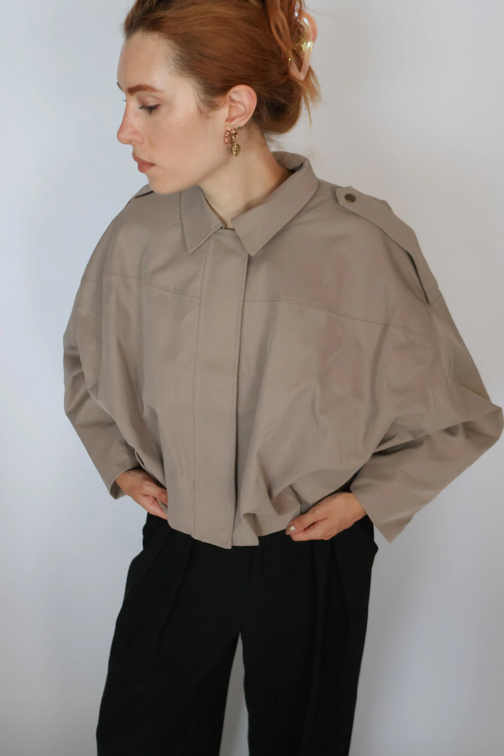 A woman wearing a cropped, oversized taupe-beige button-up shirt with structured shoulders and black trousers, standing against a plain light gray background.
