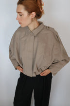 A woman wearing a cropped, oversized taupe-beige button-up shirt with structured shoulders and black trousers, standing against a plain light gray background.