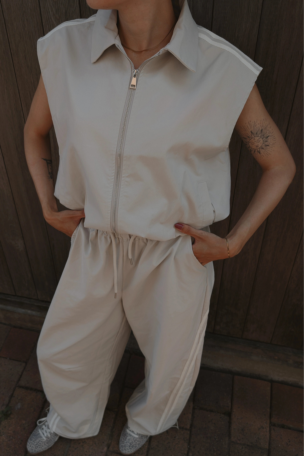 A person wearing a light beige sleeveless zip-front top and matching wide-leg drawstring pants, standing outdoors against a dark wooden fence.